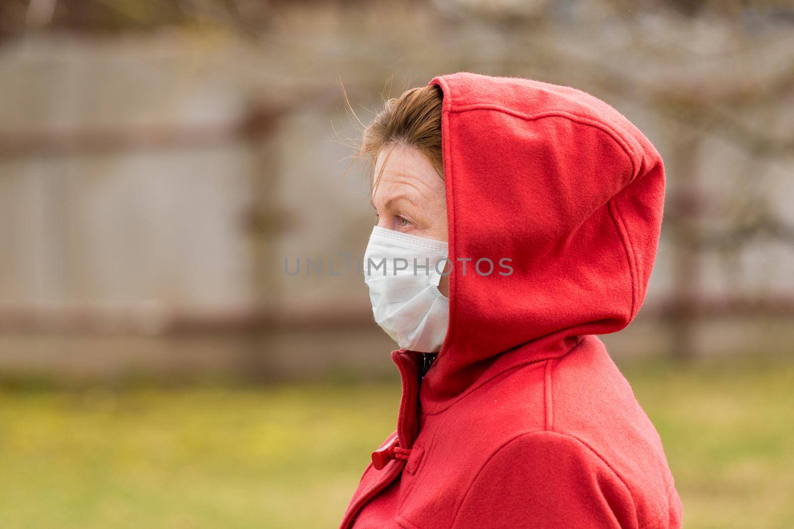 An elderly woman with brown hair and blue eyes in a red coat and hood in a protective safe medical mask, close-up portrait.