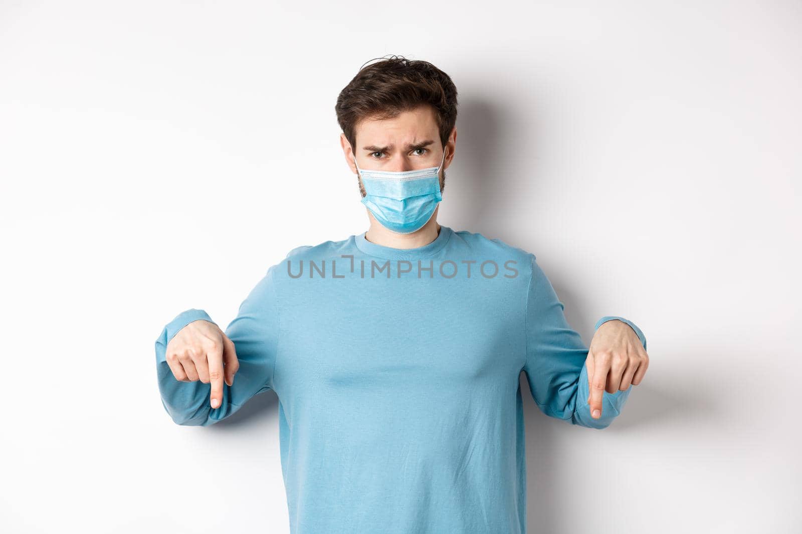 Covid-19, health and quarantine concept. Sad man complaining and looking disappointed, pointing fingers down at logo, standing in medical mask from coronavirus pandemic.