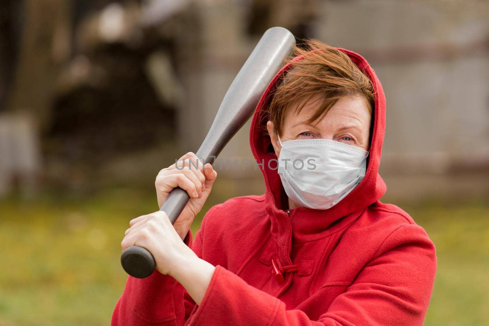 Angry aggressive elderly woman in protective safe medical mask swings baseball bat in the background of outdoor street, portrait, close up.