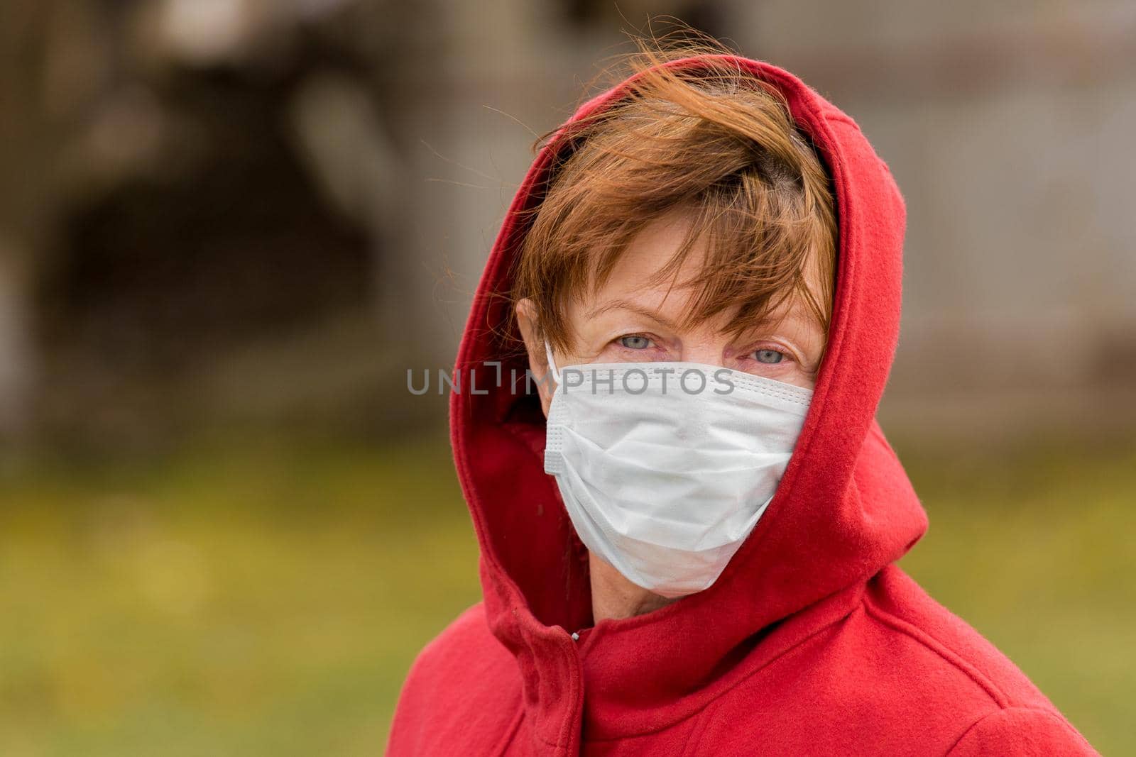 An elderly woman with brown hair and blue eyes in a red coat and hood in a protective safe medical mask, close-up portrait.