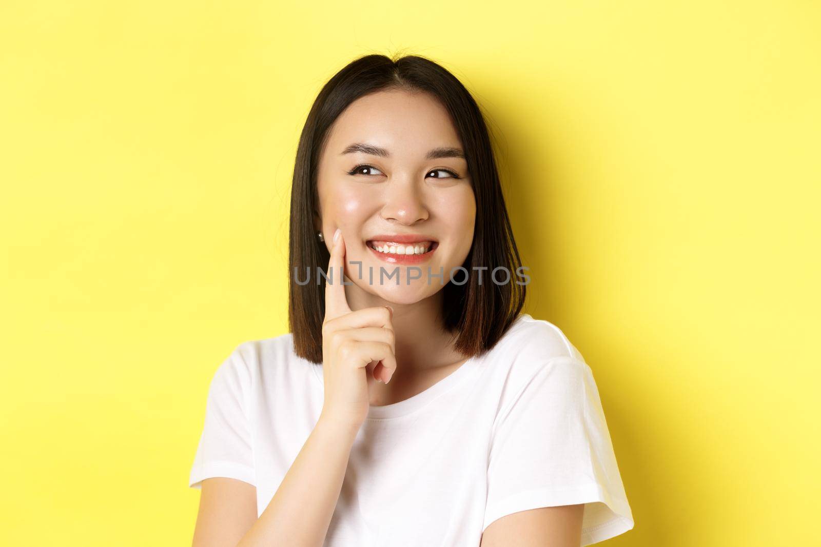 Beauty and skincare. Close up of young asian woman with short dark hair, healthy glowing skin, smiling and touching dimples on cheeks, standing over yellow background.