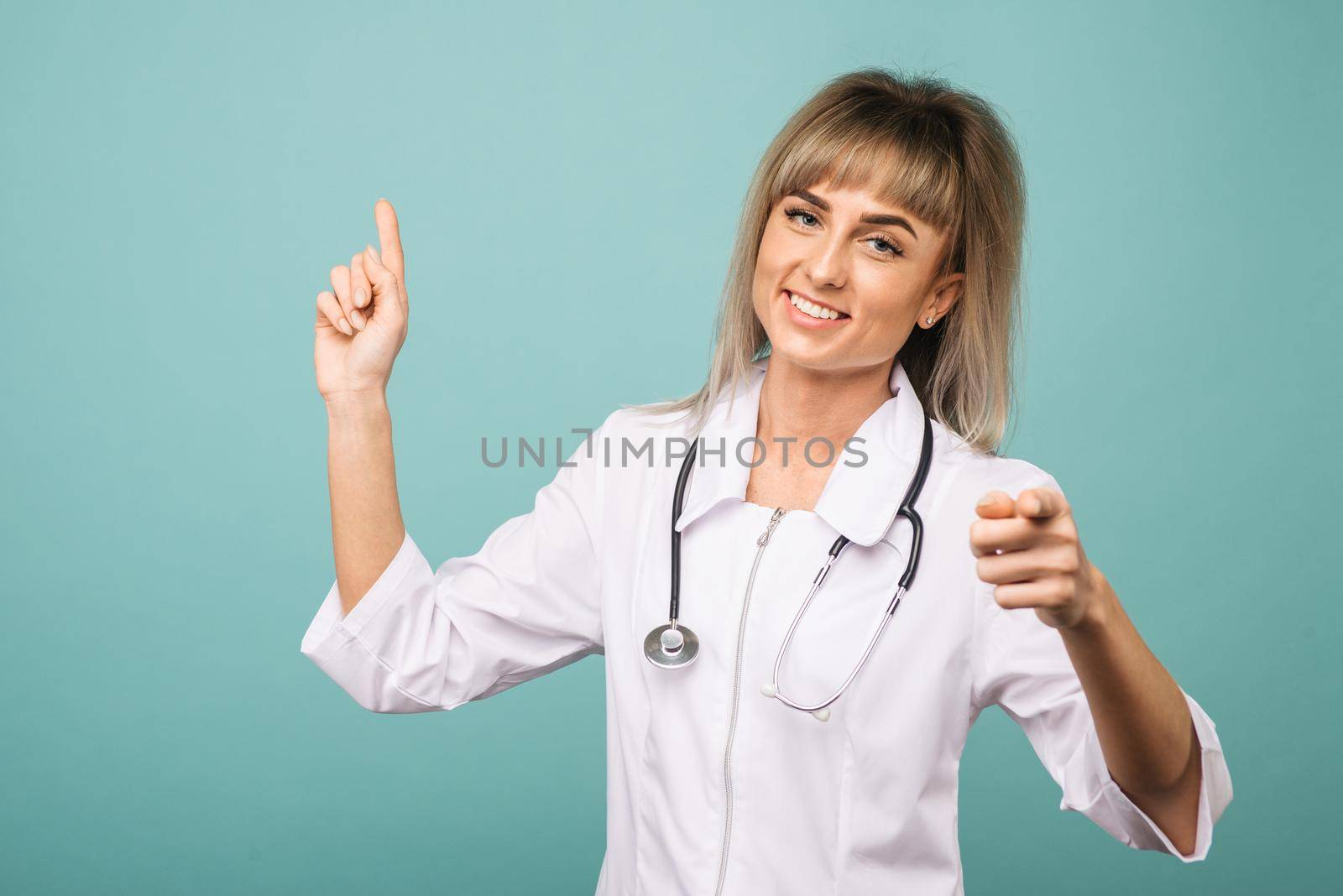Smiling young female doctor with a stethoscope points up and into the camera with her fingers on a blue background