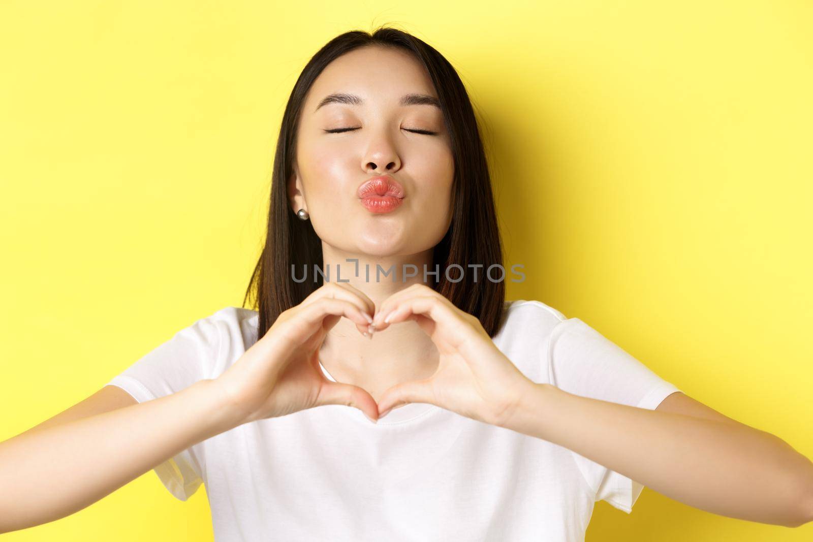 Valentines day and women concept. Close up of pretty asian girl in white t-shirt, smiling and showing heart, I love you gesture, standing over yellow background.