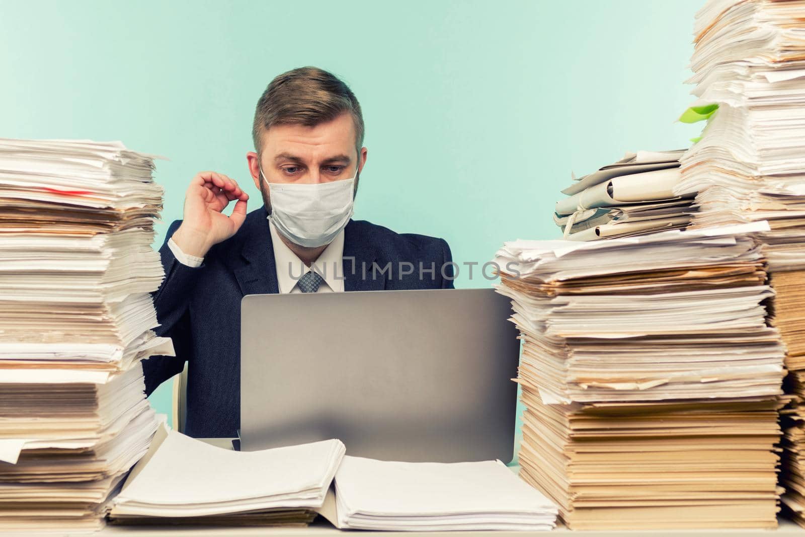 A male accountant or company manager works in an office in a pandemic in view of the accumulated paper work. A protective medical mask is on the face. On the desktop are large stacks of documents. by zartarn