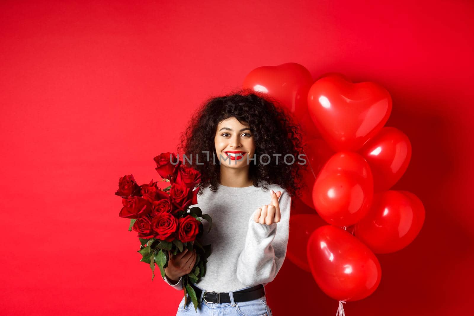 Holidays and celebration. Beautiful girlfriend receive flowers on anniversary, showing finger heart and standing near party balloons, red background.