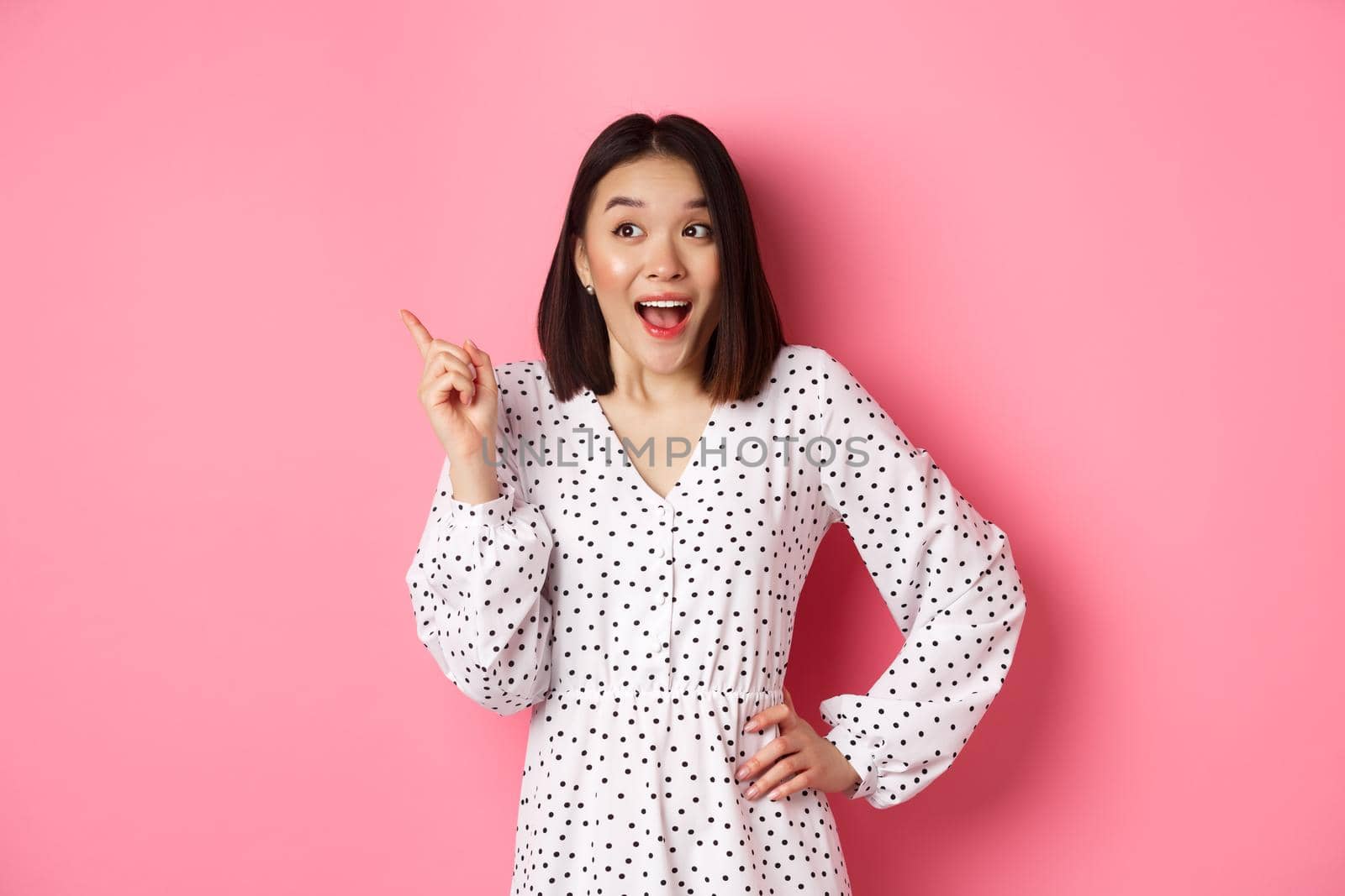 Excited asian woman in stylish dress, gasping amused, pointing finger upper left corner, showing interesting promo offer, standing over pink background.