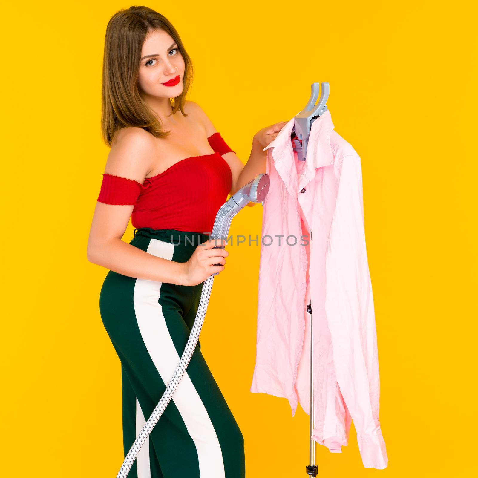 Young woman steaming pink shirt at dry-cleaner's on yellow background