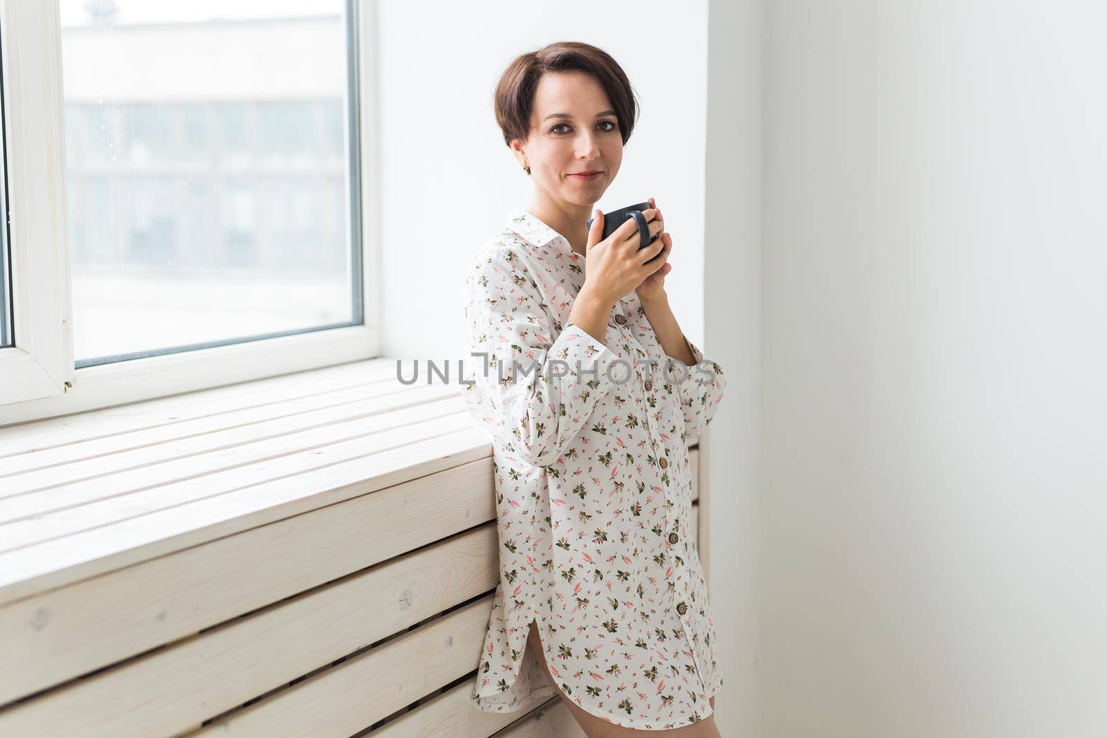 Beautiful young woman with cup of tea standing near the window at home