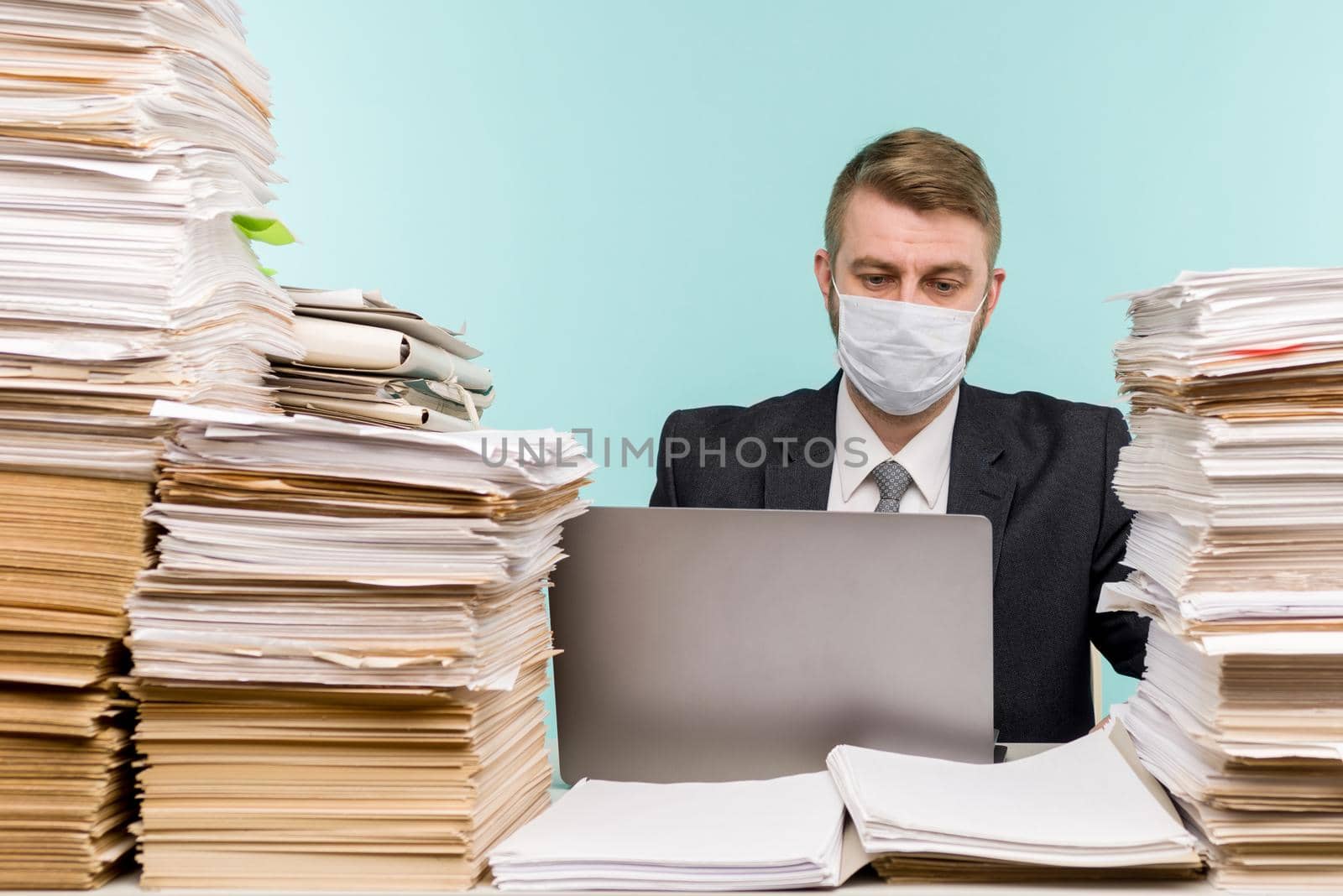 A male accountant or company manager works in an office in a pandemic in view of the accumulated paper work. A protective medical mask is on the face. On the desktop are large stacks of documents. by zartarn