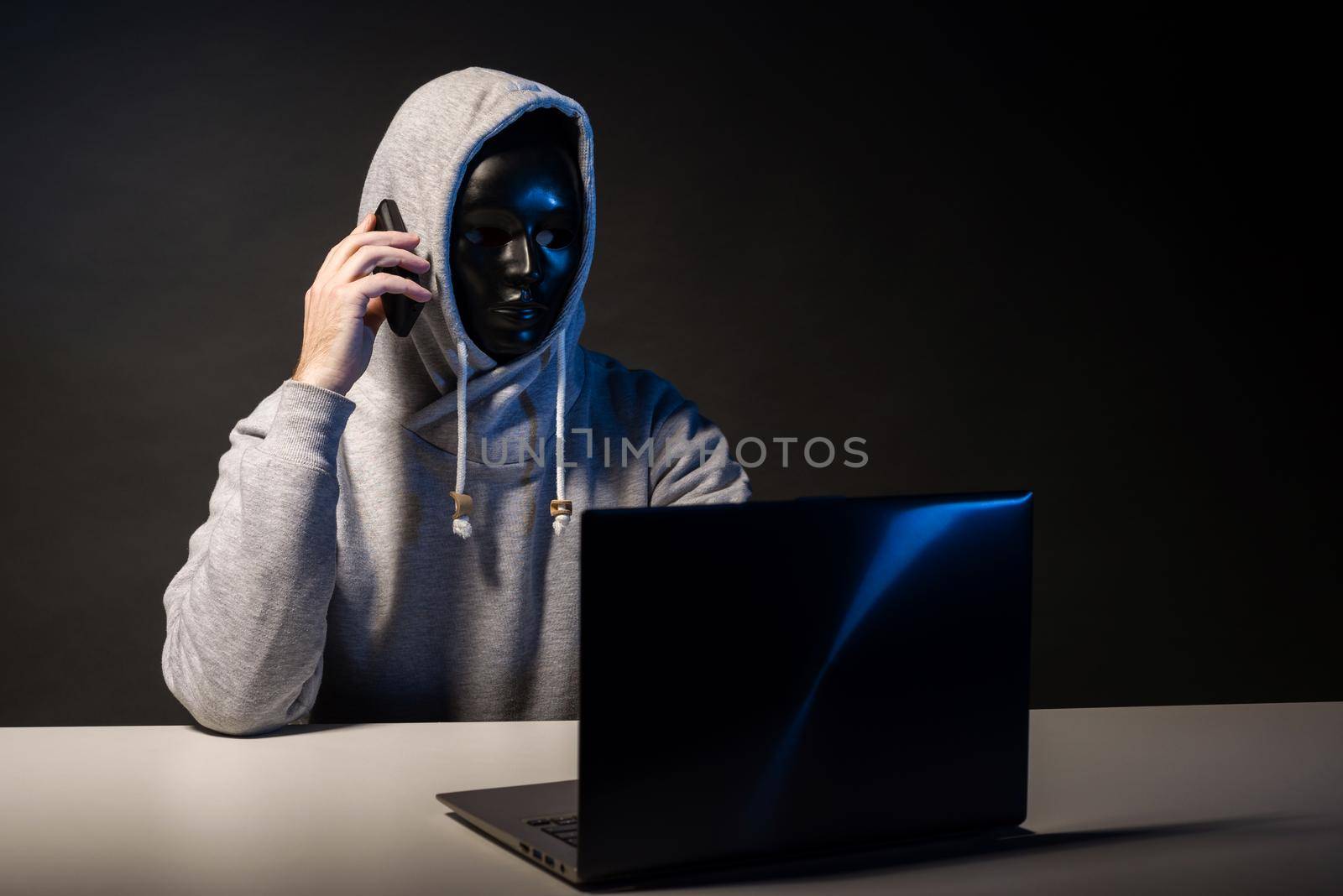 Anonymous hacker in mask programmer uses a laptop and talking on the phone to hack the system in the dark. The concept of cybercrime and hacking database