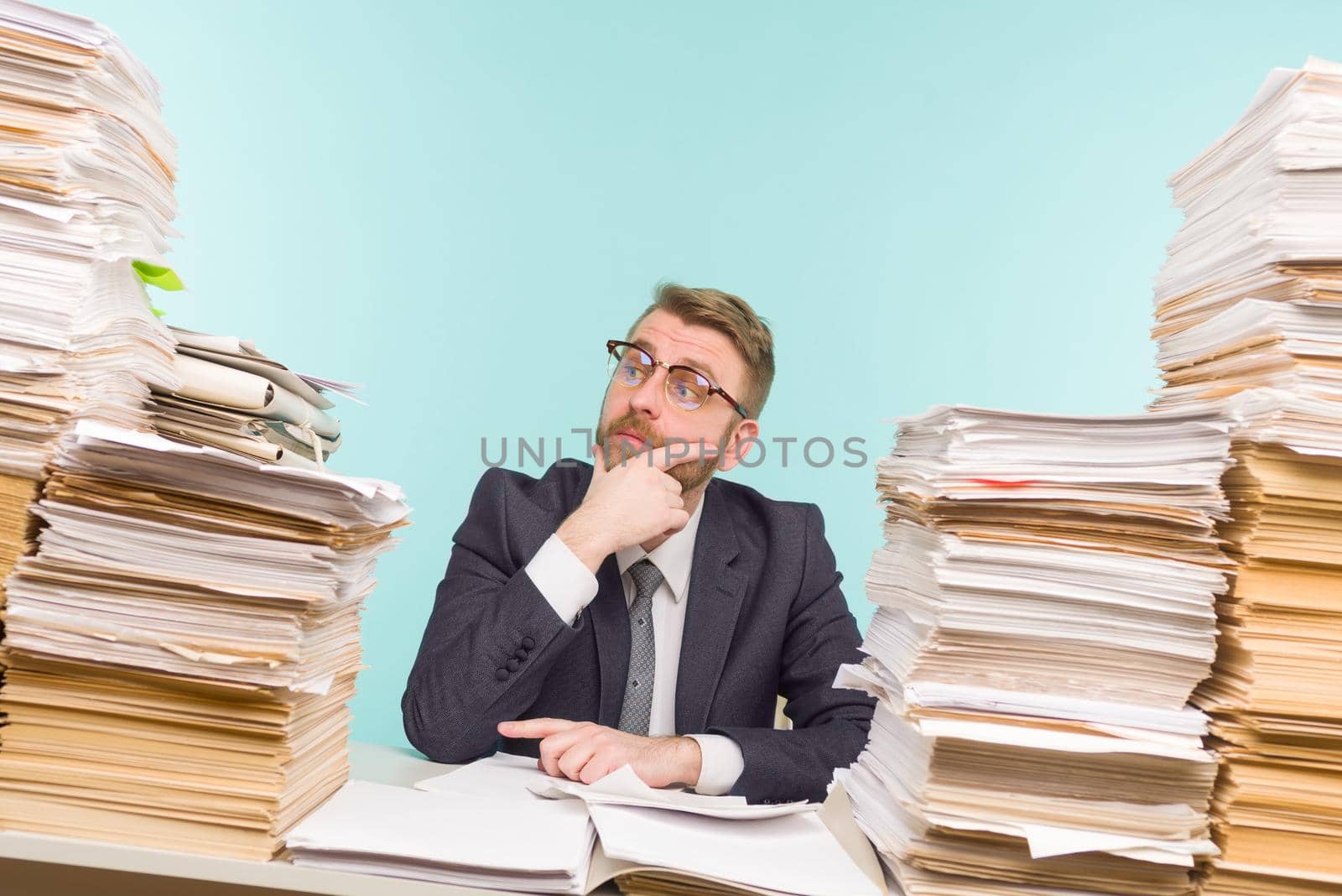 Shocked businessman sitting at the table with many papers in office, he is overloaded with work - image