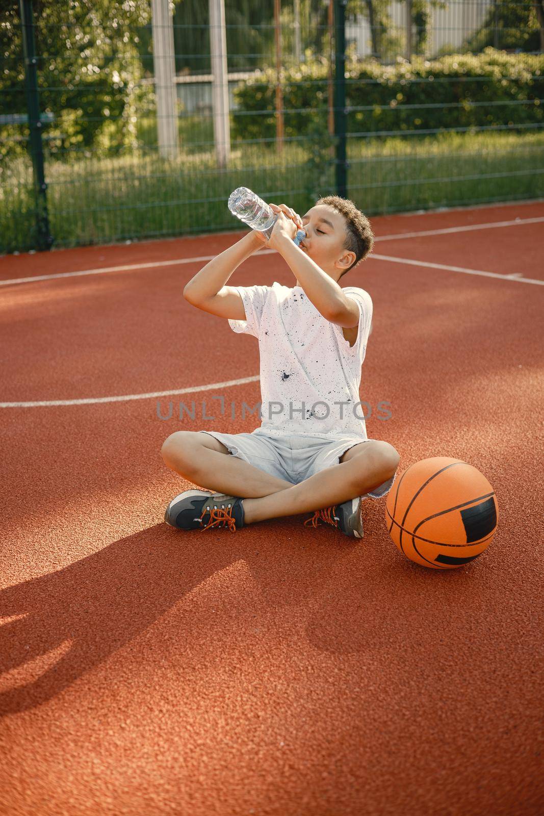 Young boy sitting on basketball court near the park. Boy wearing white t-shirt. Boy drinking a water.