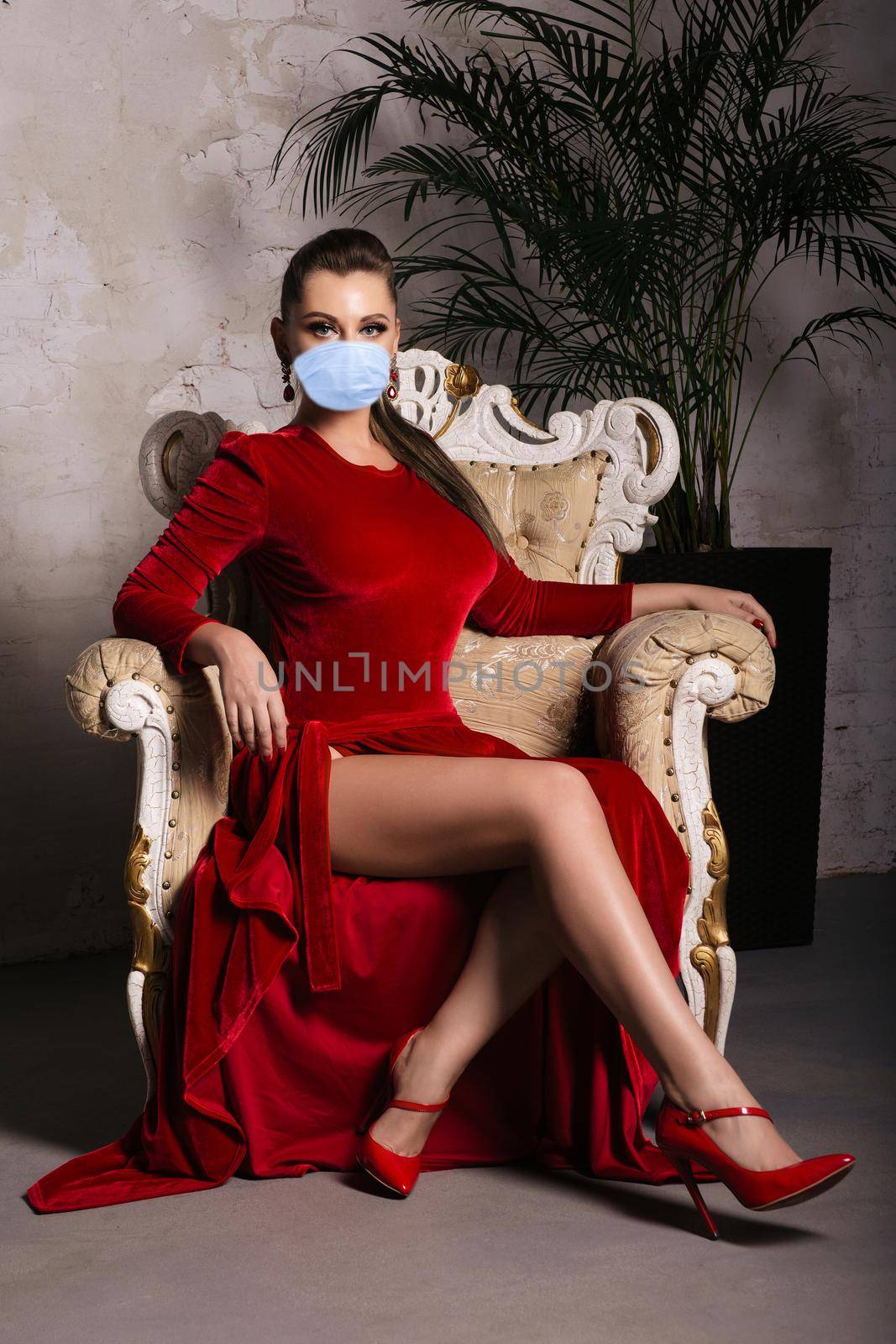 Magnificent young woman with mask in luxurious red dress and precious jewelery is sitting in a chair in a luxury apartment. Classic vintage interior. Coronavirus Outbreak Defense Concept