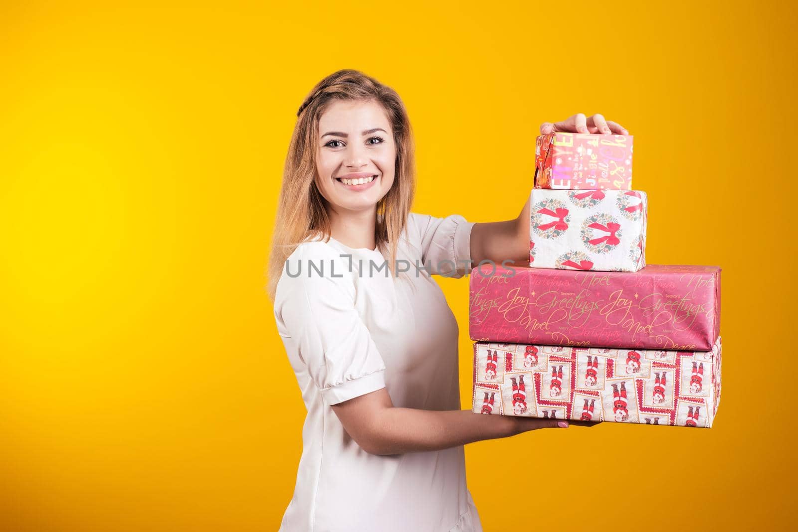 Portrait of a happy smiling girl in dress shows present boxes isolated over yellow background.