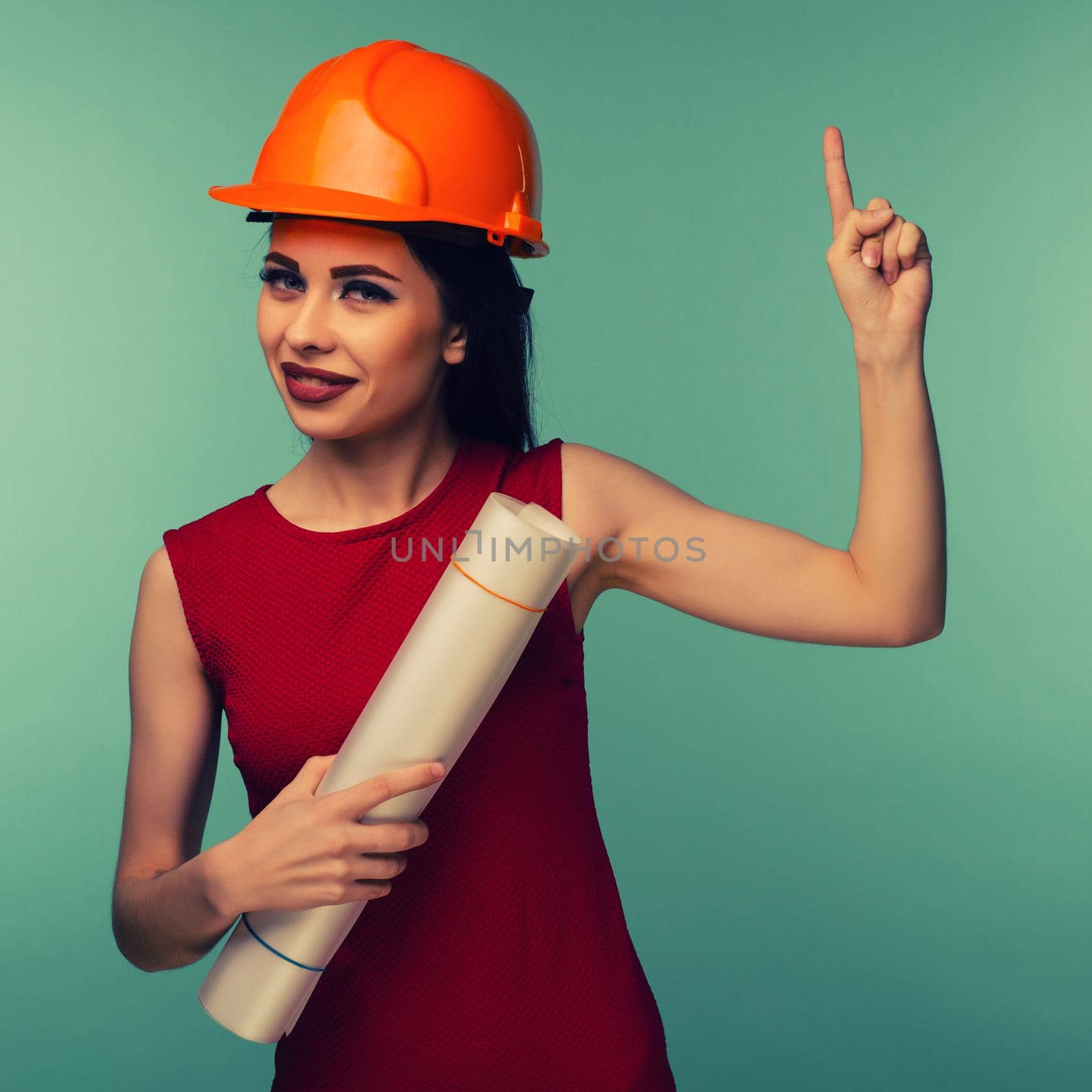 Young female engineer in orange helmet with drawings points finger up isolated on blue background - Image Toned