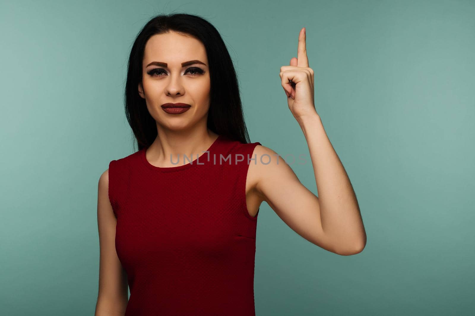 Closeup of serious strict young woman wears red dress looks stressed and pointing up with finger isolated over blue background - Image