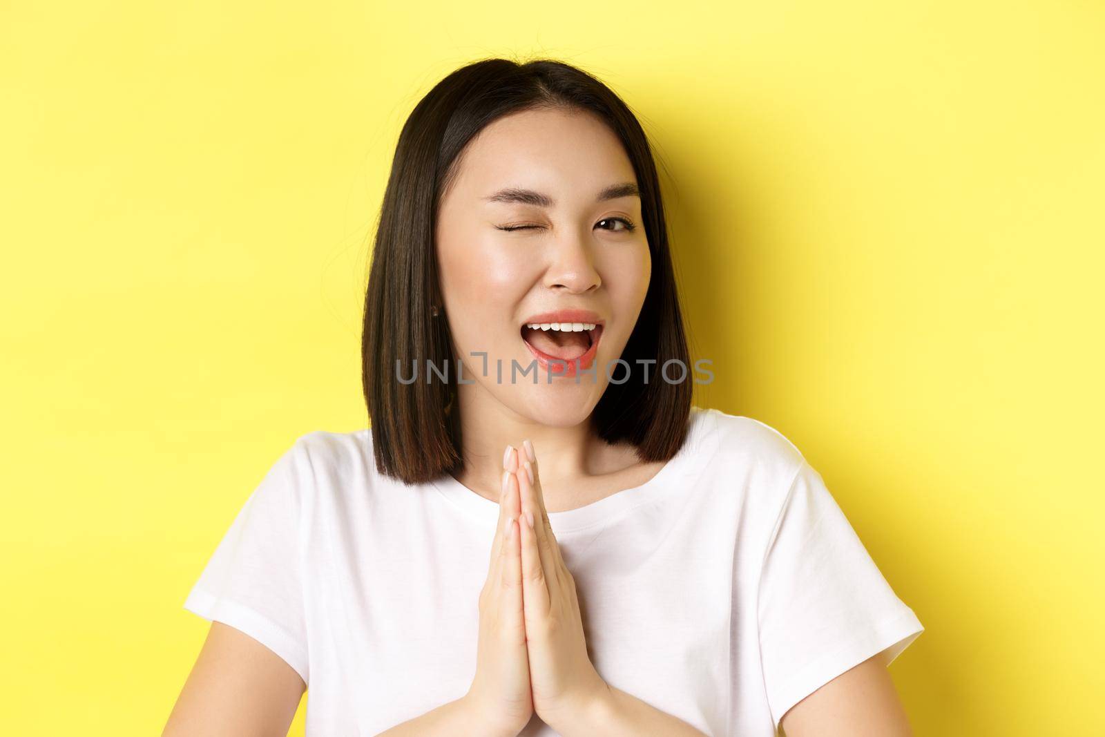 Close of cheeky young asian woman holding hands in namaste, thank you gesture, winking at camera coquettish, feeling lucky, standing over yellow background.