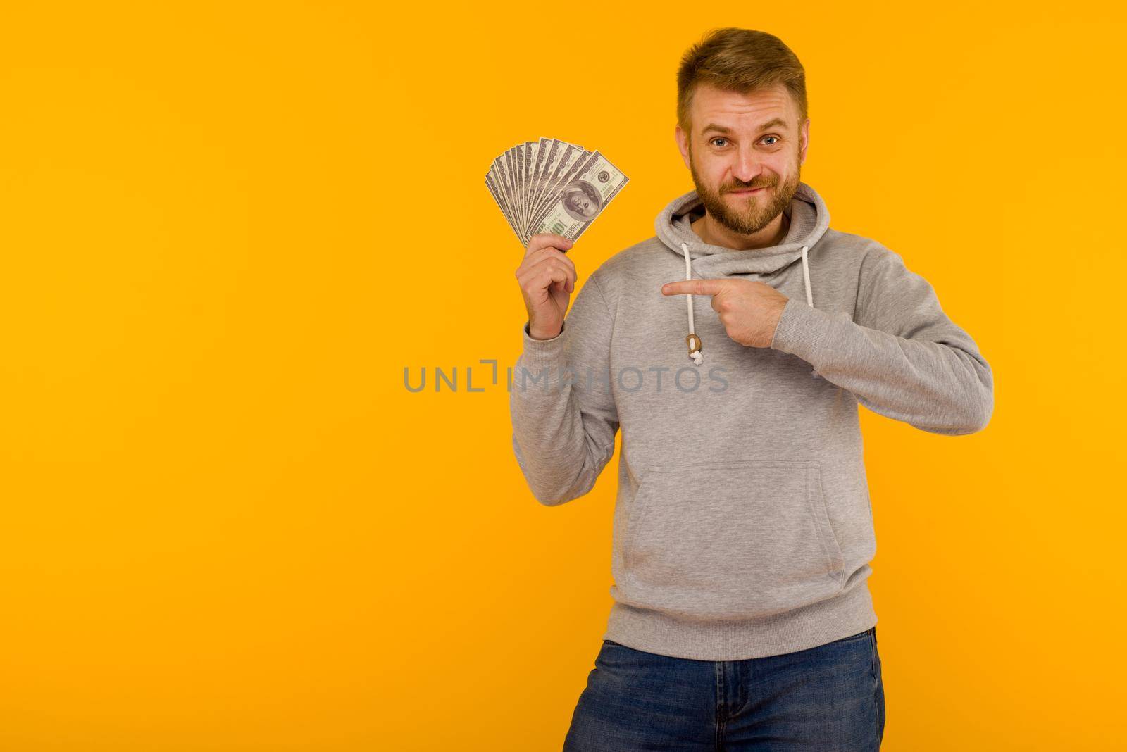 Joyful man in a gray hoodie points a finger at money dollars on a yellow background - image