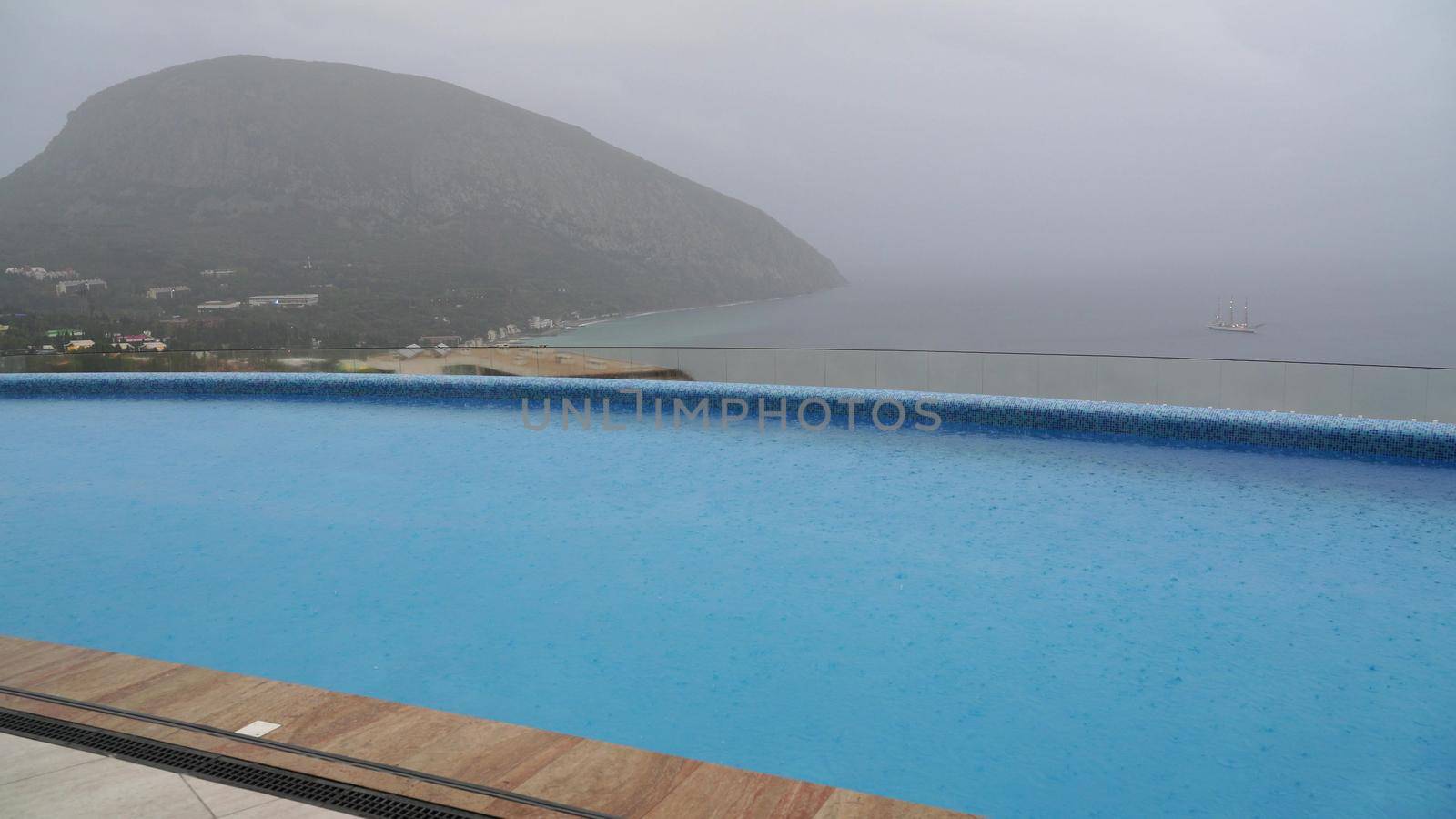 Autumn rain falls in large drops on the heated outdoor pool. Swimming pool in the hotel on the background of the mountain and the sea. by Matiunina