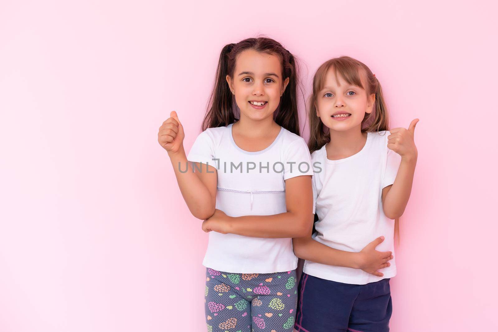 Children showing like. Two adorable happy little girls gesturing thumbs up together and smiling to camera, excellent feedback, good job. indoor studio shot isolated on pink background