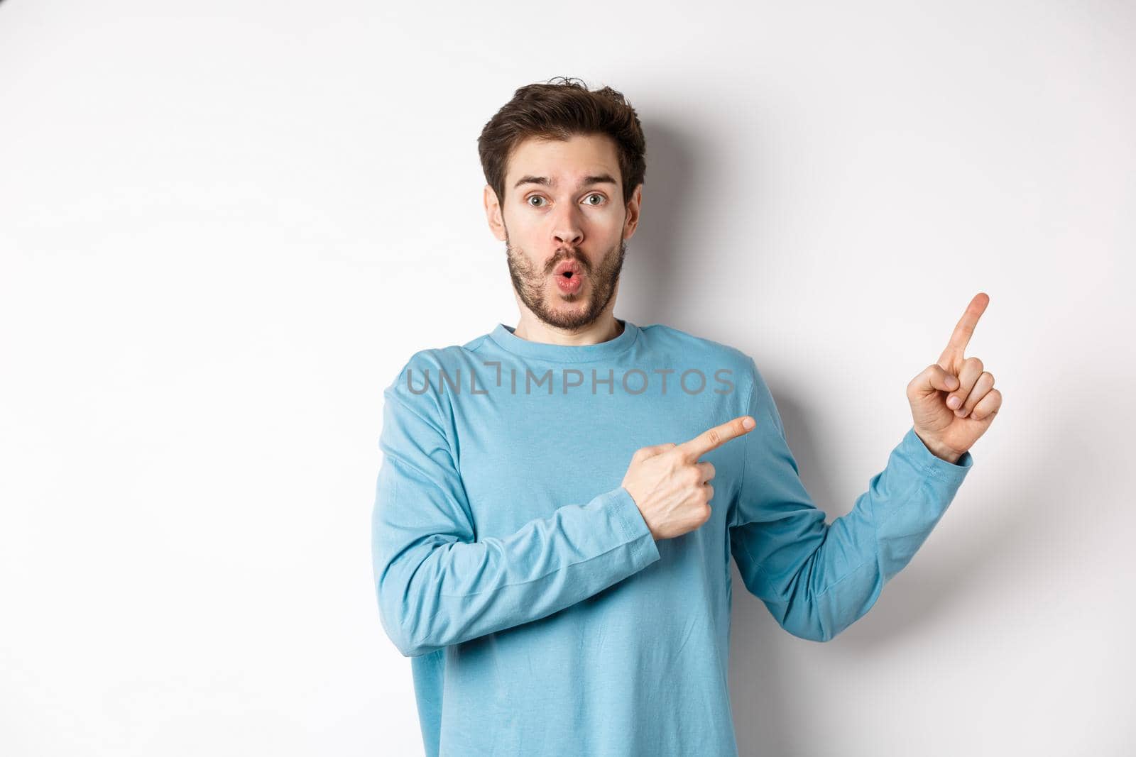 Wow look here. Impressed young man pointing fingers right and showing logo, standing amazed on white background.