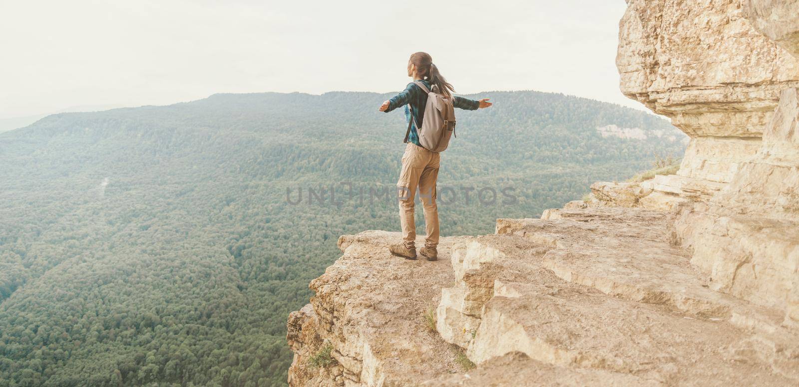 Backpacker young woman standing on cliff Eagle shelf with raised arms and enjoying view of nature, Mezmay, Krasnodar region, Russia