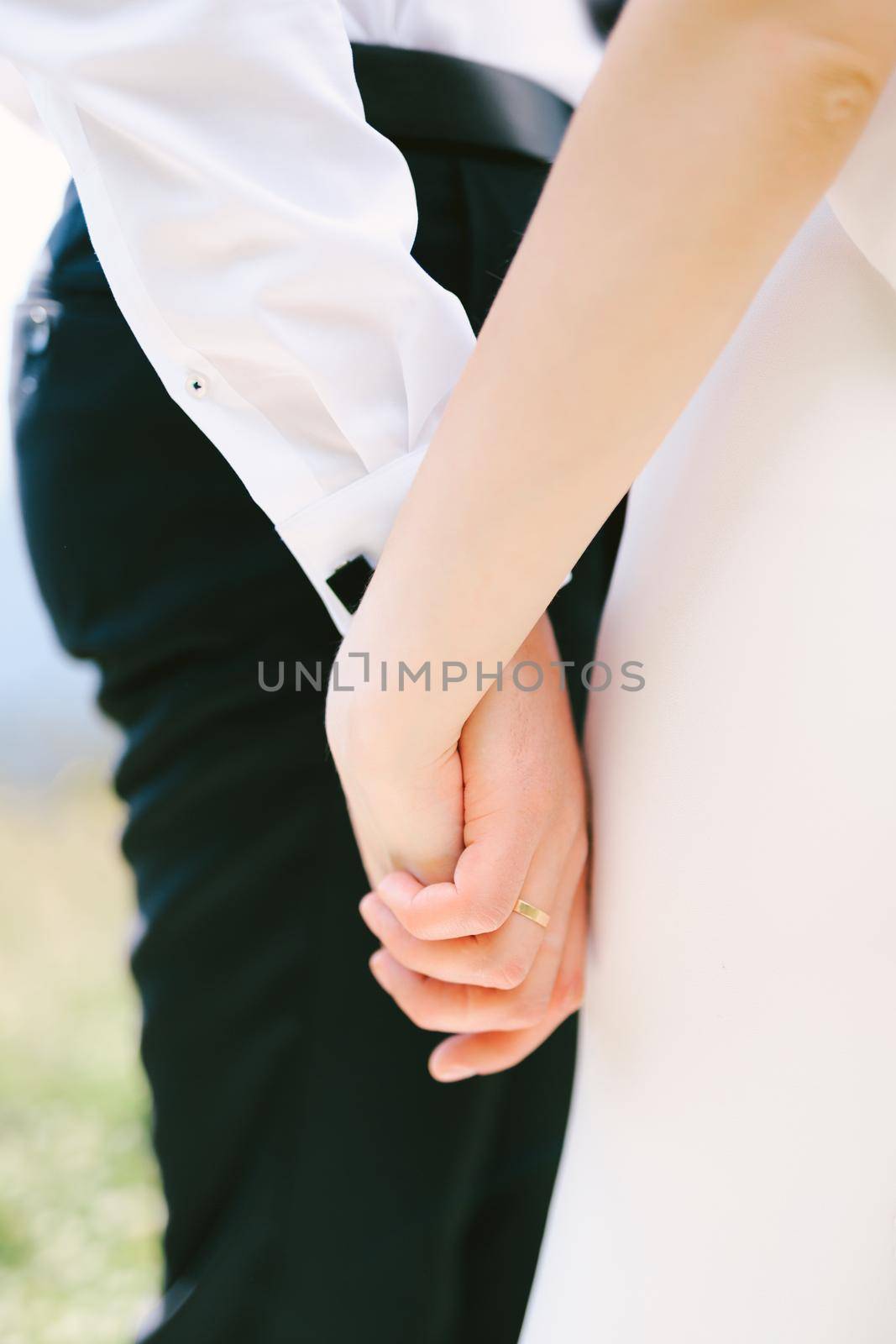 Hand of the groom in a white shirt and black trousers holds the hand of bride in a white dress. Close-up. High quality photo