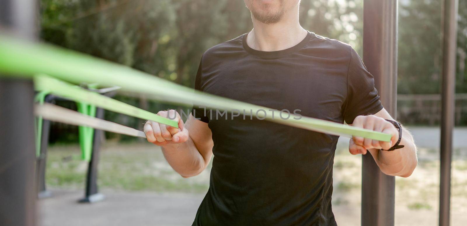 Strong man guy training with elastic rubber band at the stadium outdoors. Adult male person during workout for arms and chest with additional sport equipmant. Sportsman exercising outside