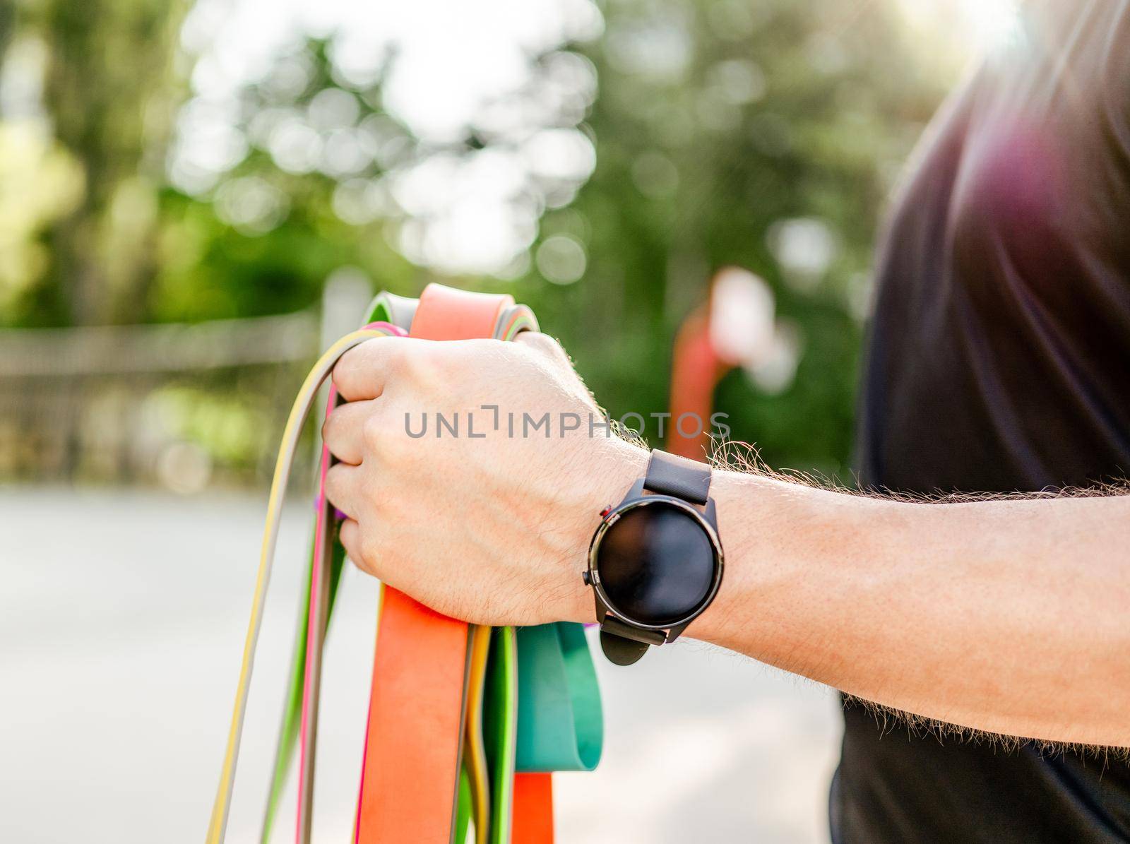 Strong man guy holding in his hand wearing fitness tracker set of colorful elastic rubber bands at the stadium outdoors. Athlete male person during workout with additional sport equipmant