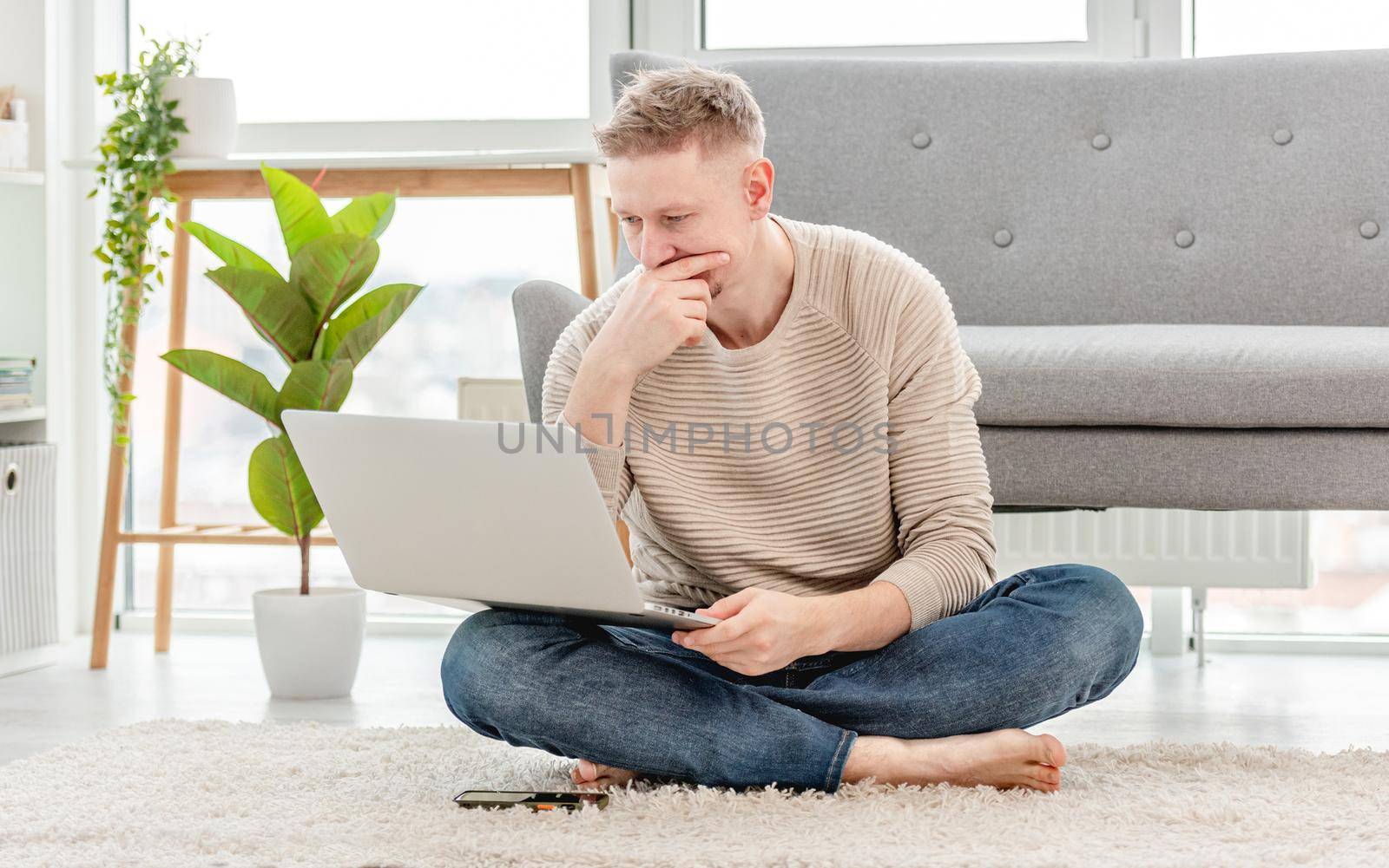 Man freelancer looking at laptop and thinking during remote work time at home. Adult guy sitting on the floor searching in internet