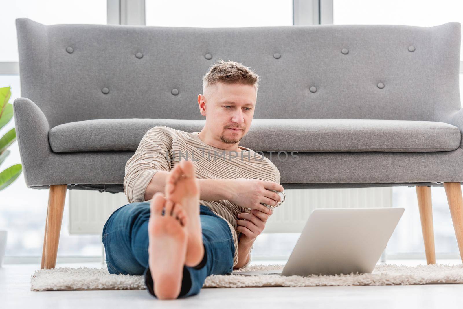 Man sitting on the floor with coffee and reading in laptop at home. Light beige interior workspace