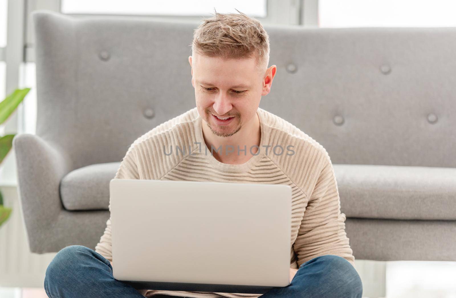 Attractive blond man sitting with laptop, typing and chatting using internet. Home workspace during remote daily work