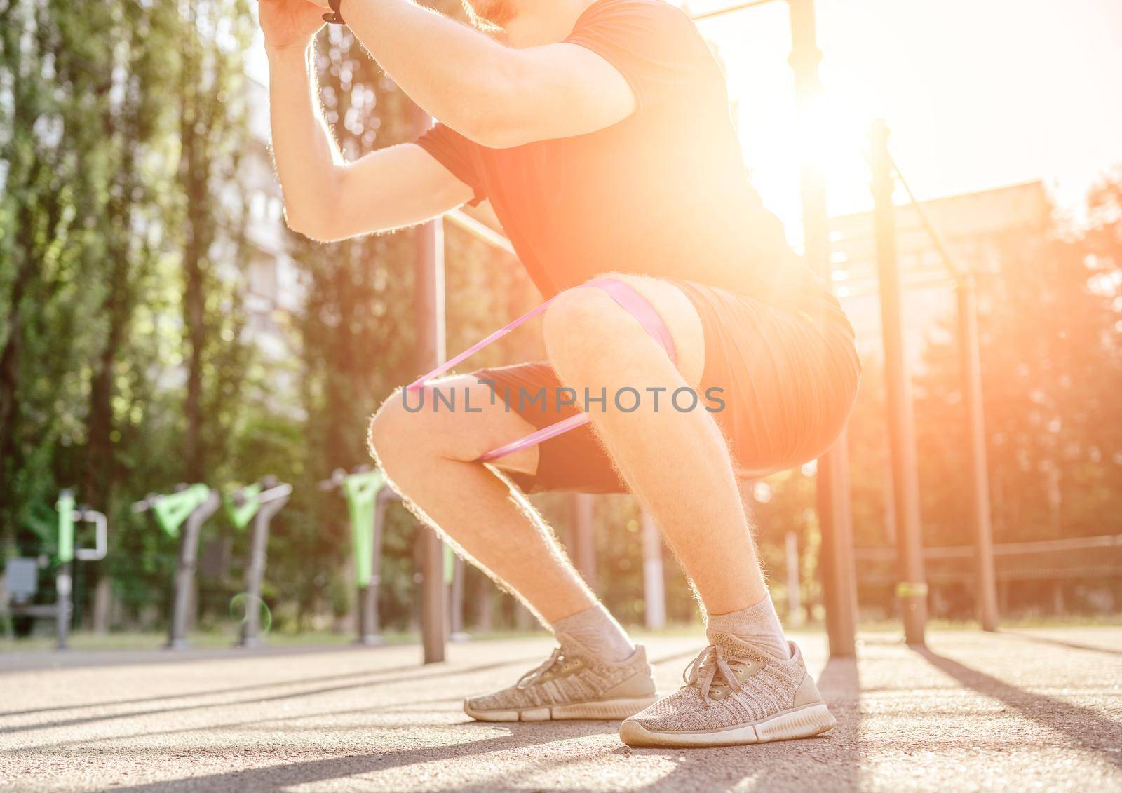 Strong sportive man guy doing squats with elastic rubber band at the stadium outdoors with sunlight. Adult male person during workout for legs with additional sport equipmant