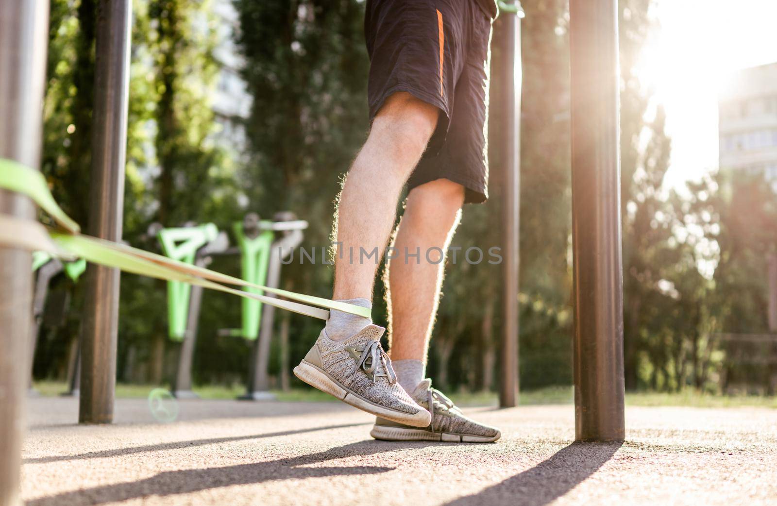 Strong man guy training with elastic rubber band at the stadium outdoors. Adult male person during workout for legs with additional sport equipmant. Sportsman exercising outside