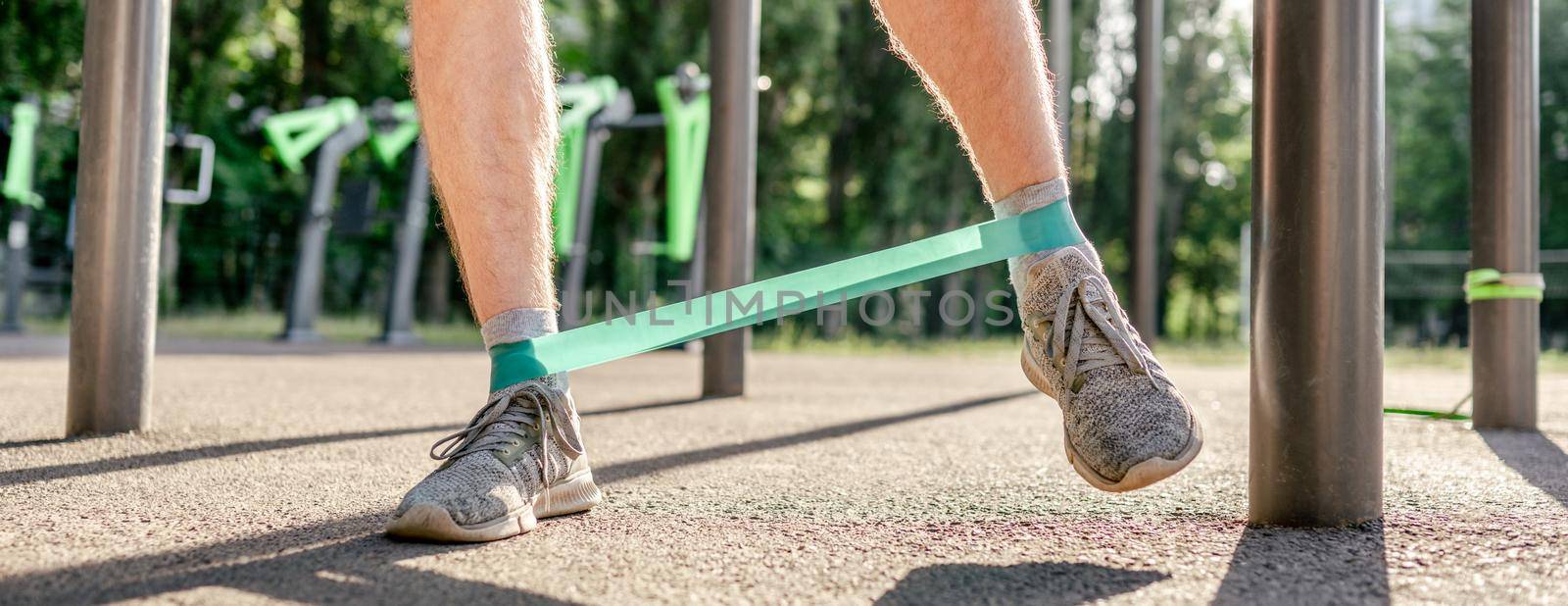 Closeup front view of legs feets of man guy training with elastic rubber band at the stadium outdoors. Athlete adult male person during active workout with additional sport equipmant in summertime