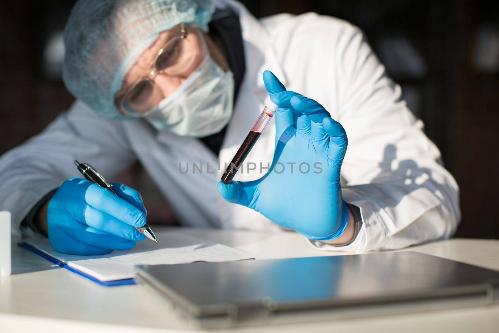 Close-up of a doctor in protective mask and wear holding a test tube with a sample of infected blood and make a notes. by Stavros