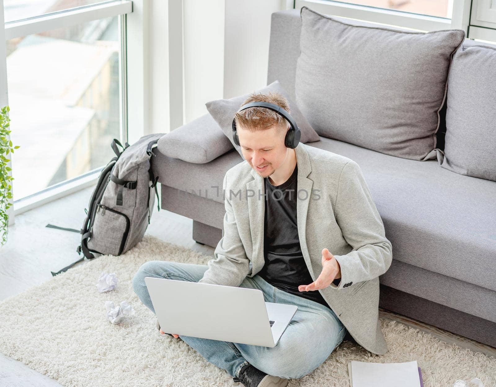 Cheerful man talking online on computer sitting on floor in light apartment
