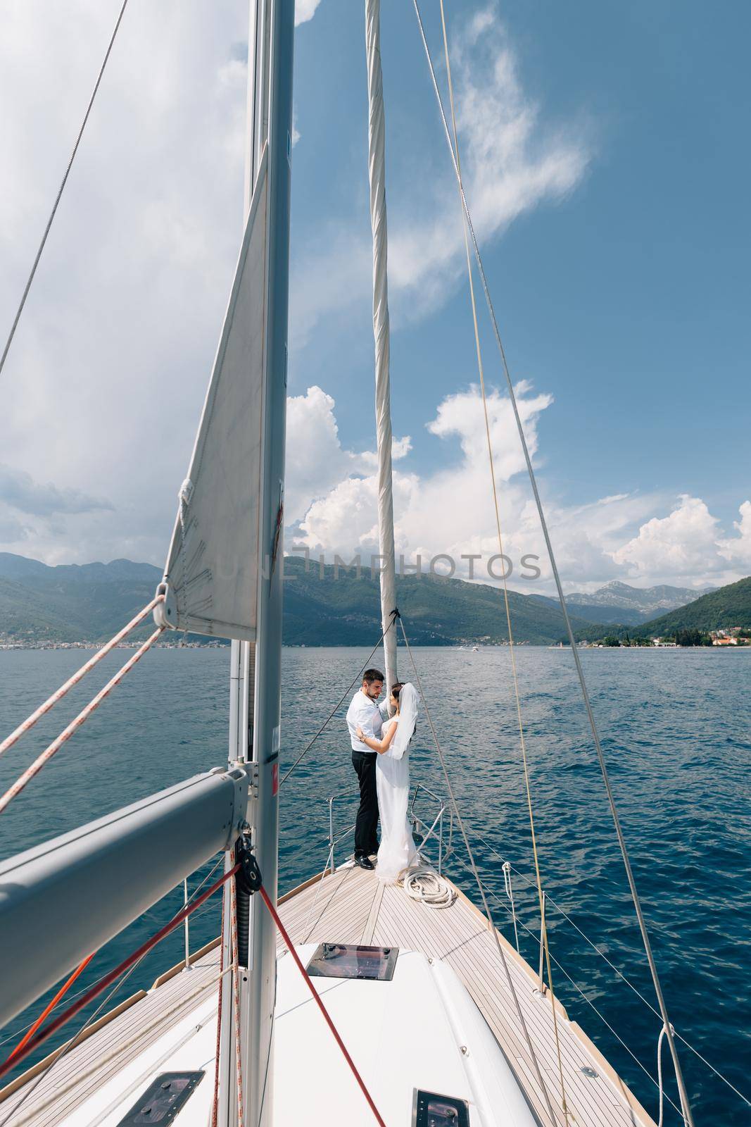 The bride and groom are hugging while standing on the bow of a white yacht sailing in the Bay of Kotor by Nadtochiy