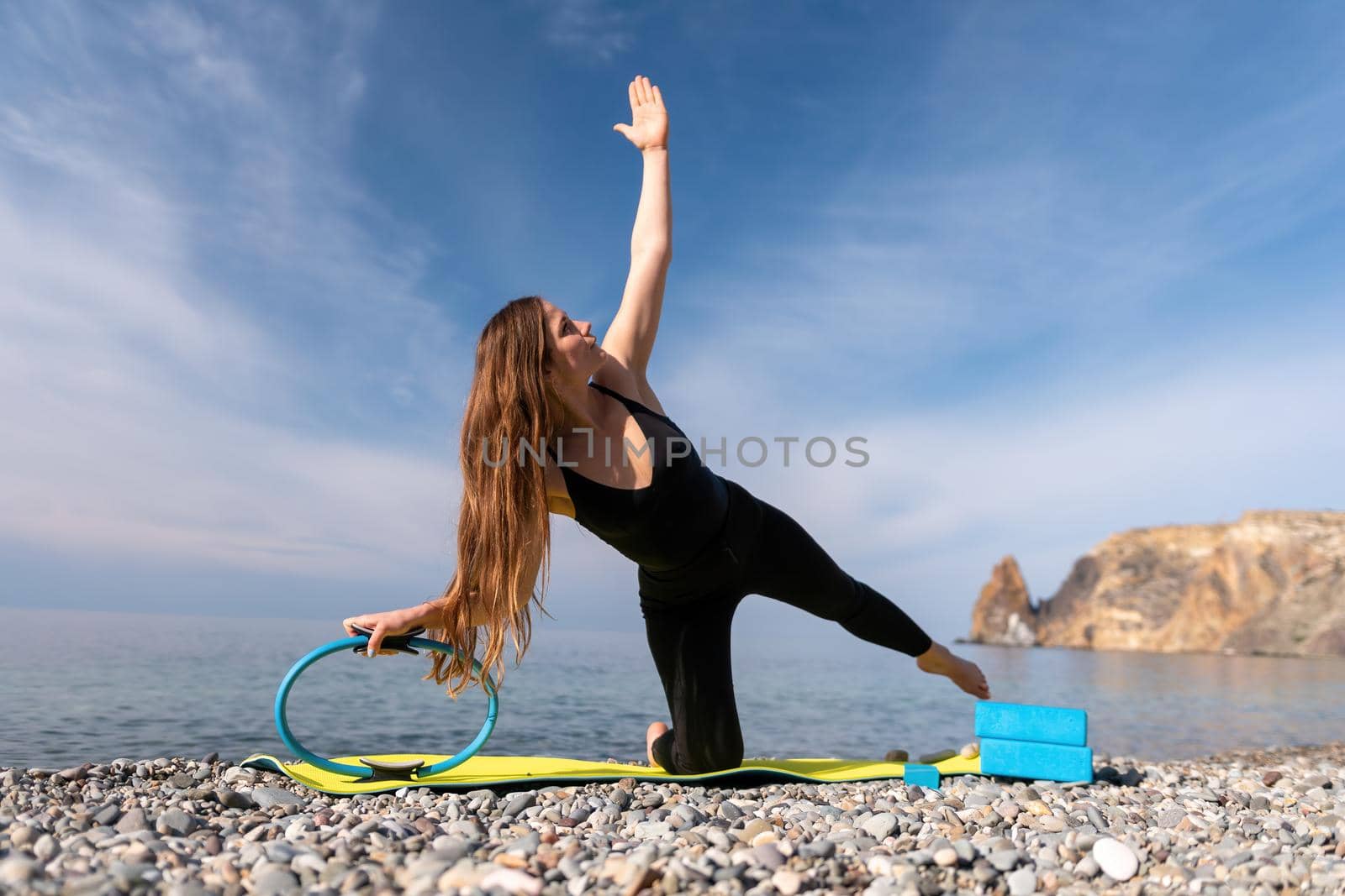 Young woman with long hair, fitness instructor in black Sportswear Leggings and Tops, stretching on a yoga mat with magic pilates ring near the sea on a sunny day, female fitness yoga routine concept by panophotograph