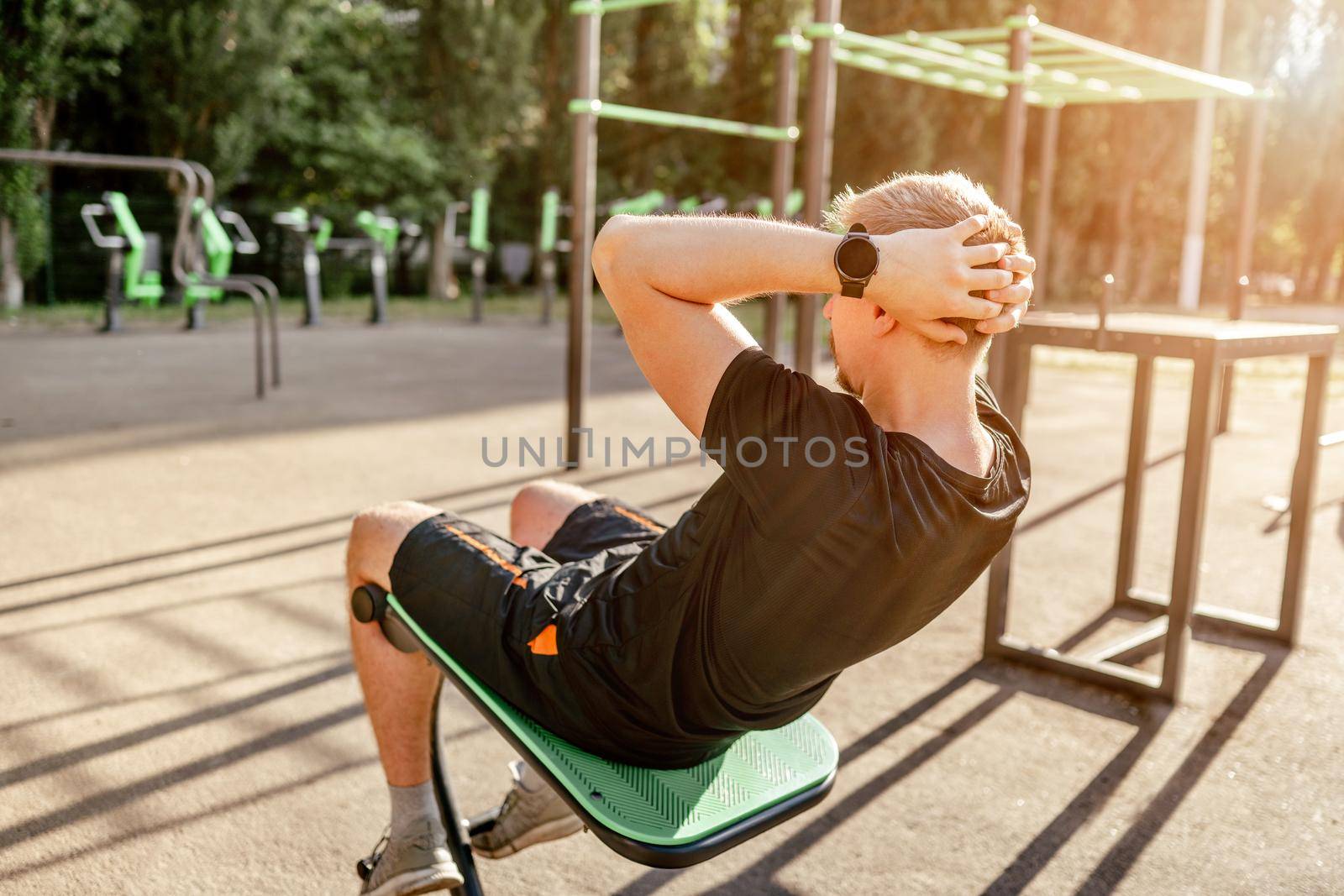 Strong sportive man guy pumps his abs at the stadium outdoors. Adult male person during workout for tummy with additional sport equipmant. Sportsman exercising outside