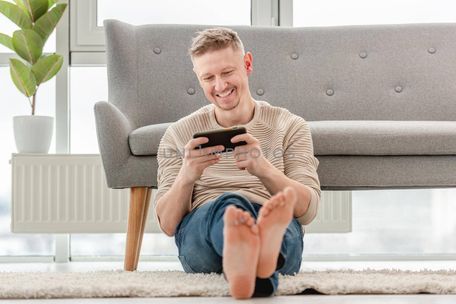 Attractive blong man playing games on smartphone, sitting on the floor and smiling. Online global gaming connection