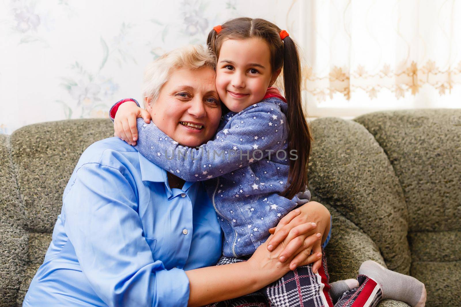 Little cute girl and her grandmother are spending time together at home. Having fun, hugging and smiling while sitting on sofa. by Andelov13