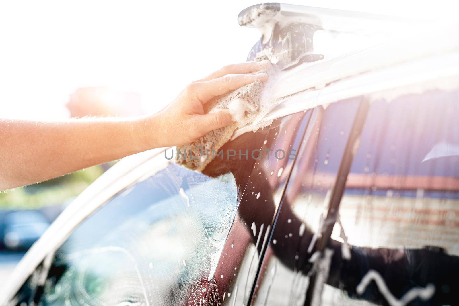 Hand with sponge is cleaning car under sparkling sun