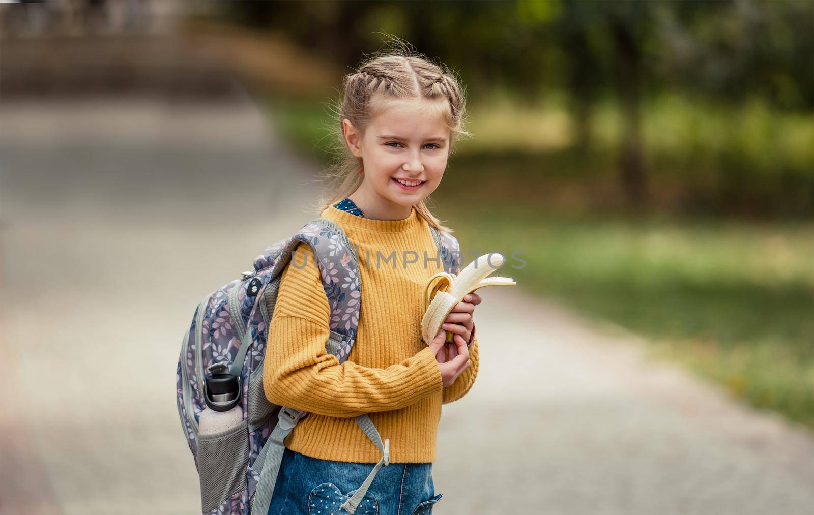 Preteen school girl with backpack holding bananas in her hands, looking at camera and smiling. Pretty pupil female kid after education class