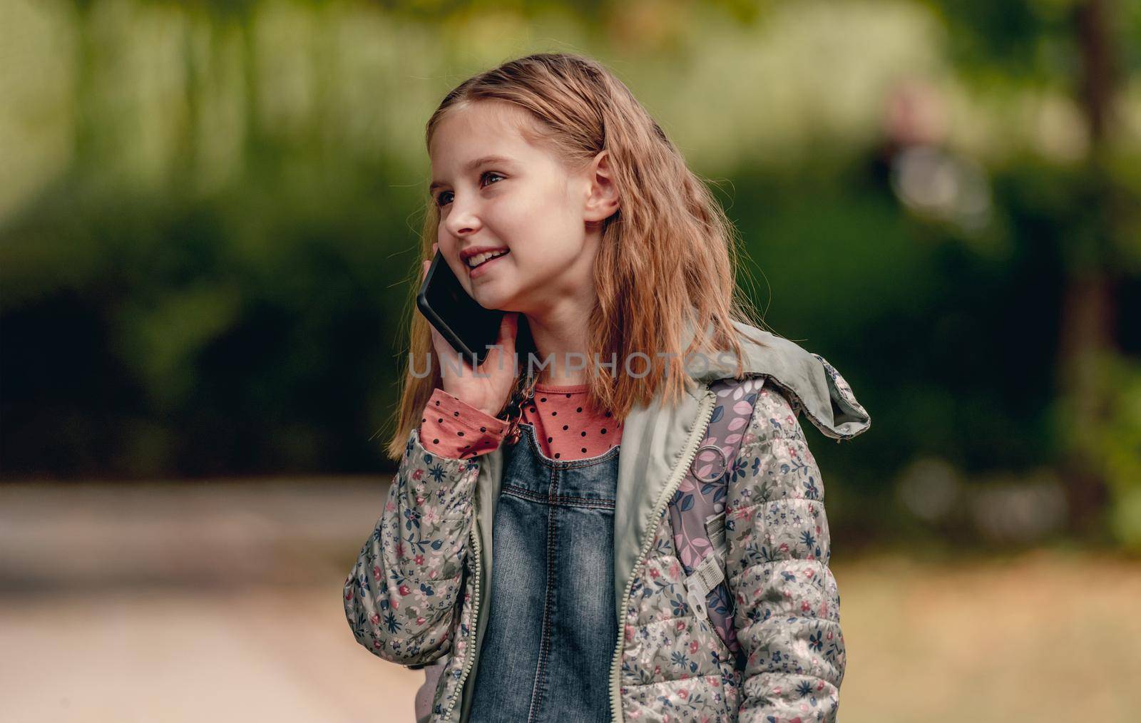 Pretty school girl talking by smartphone outdoors and smiling. Cute female kid communicates using cell phone in autumn park