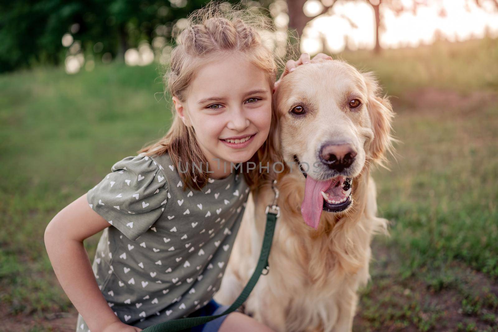 Portrait of beautiful preteen girl with golden retriever dog looking at camera together outdoors. Kid with doggy pet in the field in summer time