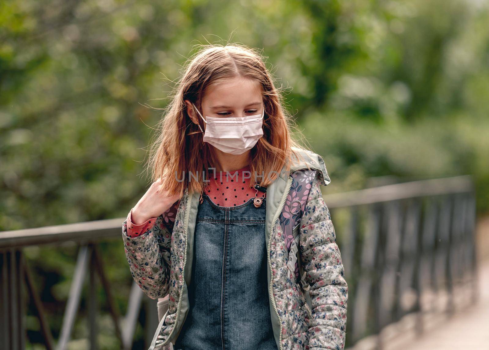 Pretty school girl wearing face mask standing in autumn park and looking at camera. Beautiful female kid after school in pandemic coronavirus covid time