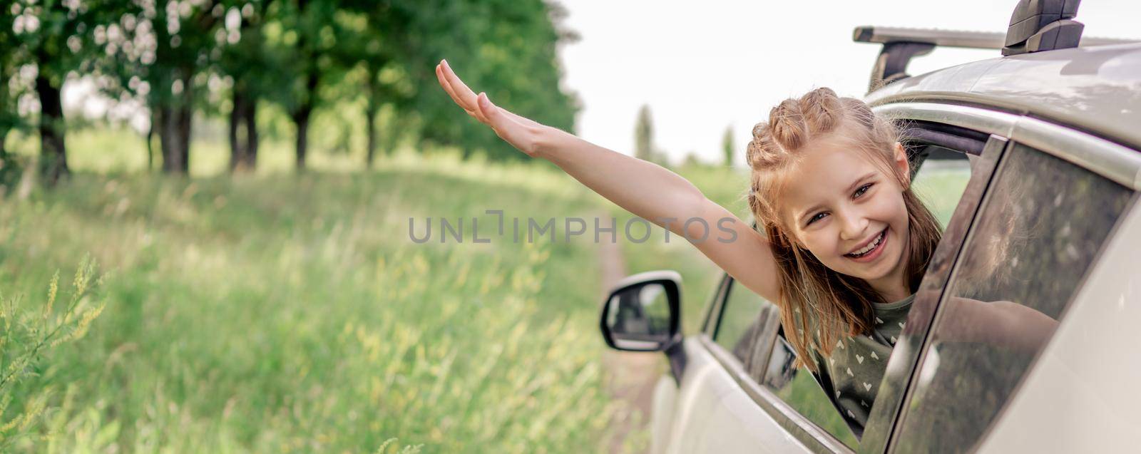 Adorable preteen girl sitting in the car and looking out the window open and smiling. Portrait of happy child kid in the vehicle outdoors in the field during summer trip