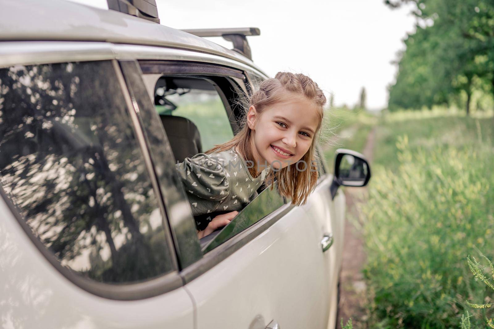 Beautiful preteen girl sitting in the car and looking out the window open and smiling in the field. Child kid in the vehicle outdoors during summer journey