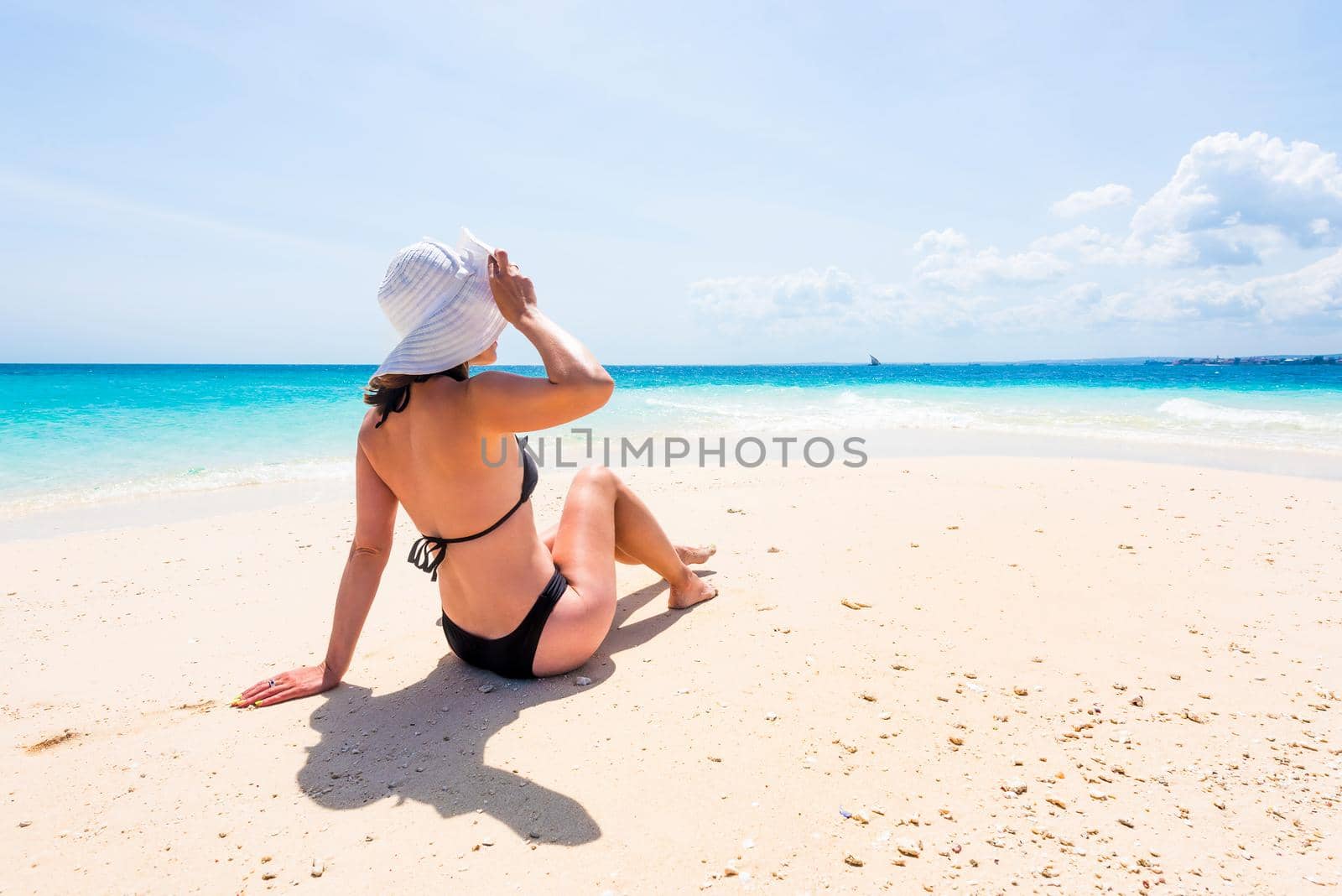 girl in hat and black swimsuit sitting on beautiful beach with blue ocean and sky on the background